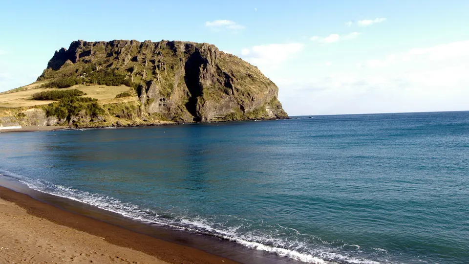 A large, rugged cliff face covered in green vegetation rises from the sea, with a sandy beach in the foreground.