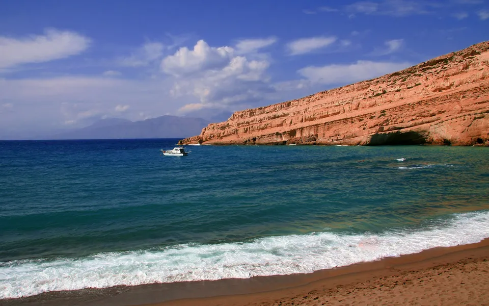 A small white boat floats in the turquoise sea near a sandy beach and dramatic red cliffs under a blue sky with clouds.
