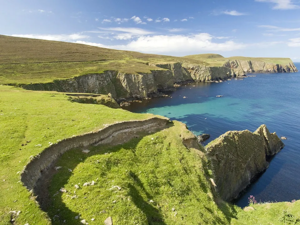 Green grassy cliffs drop sharply to the clear blue sea, with a rugged rock formation in the foreground.