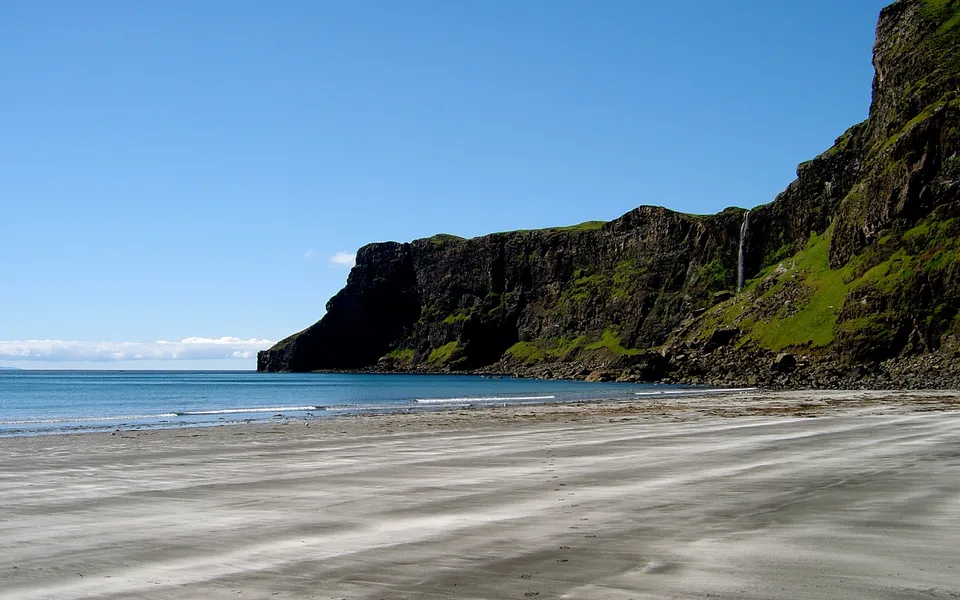 A wide sandy beach leads to the ocean, with dramatic, green-covered cliffs and a waterfall cascading down.