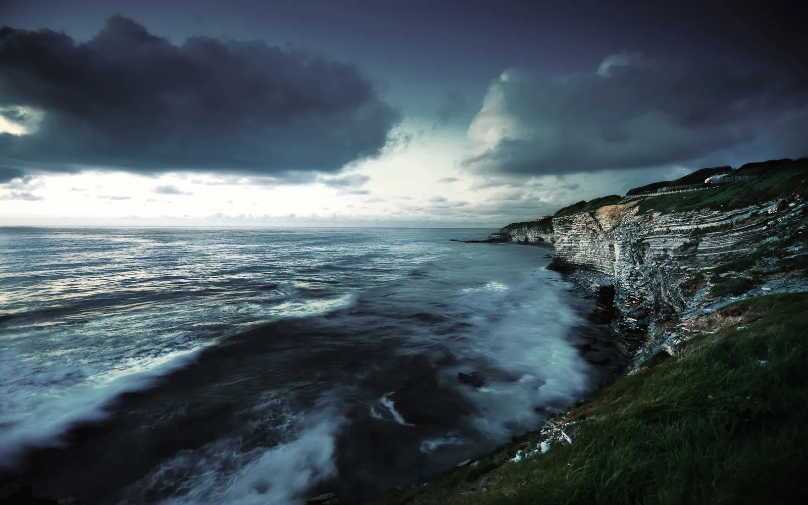 Waves crash against layered cliffs under a dramatic, cloudy sky.