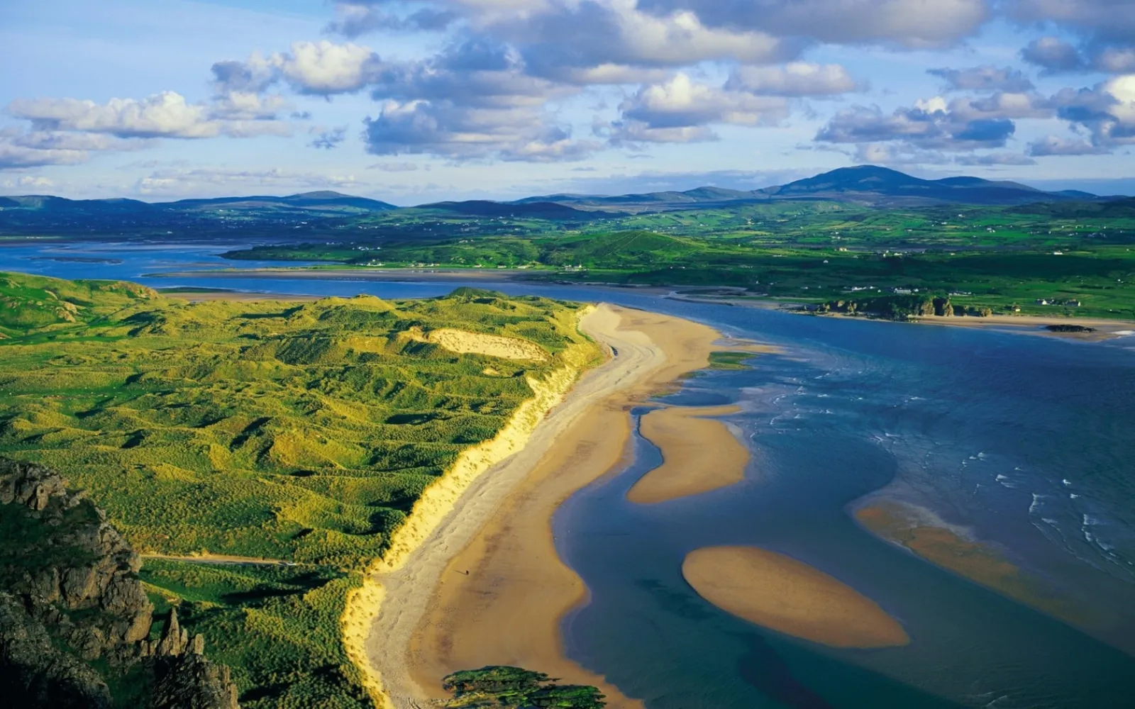 An aerial view of a coastline with rolling green dunes meeting a wide expanse of blue water and a sandy beach.