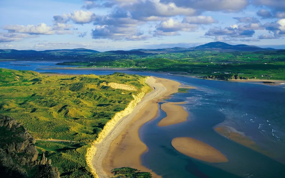 An aerial view of a coastline with rolling green dunes meeting a wide expanse of blue water and a sandy beach.