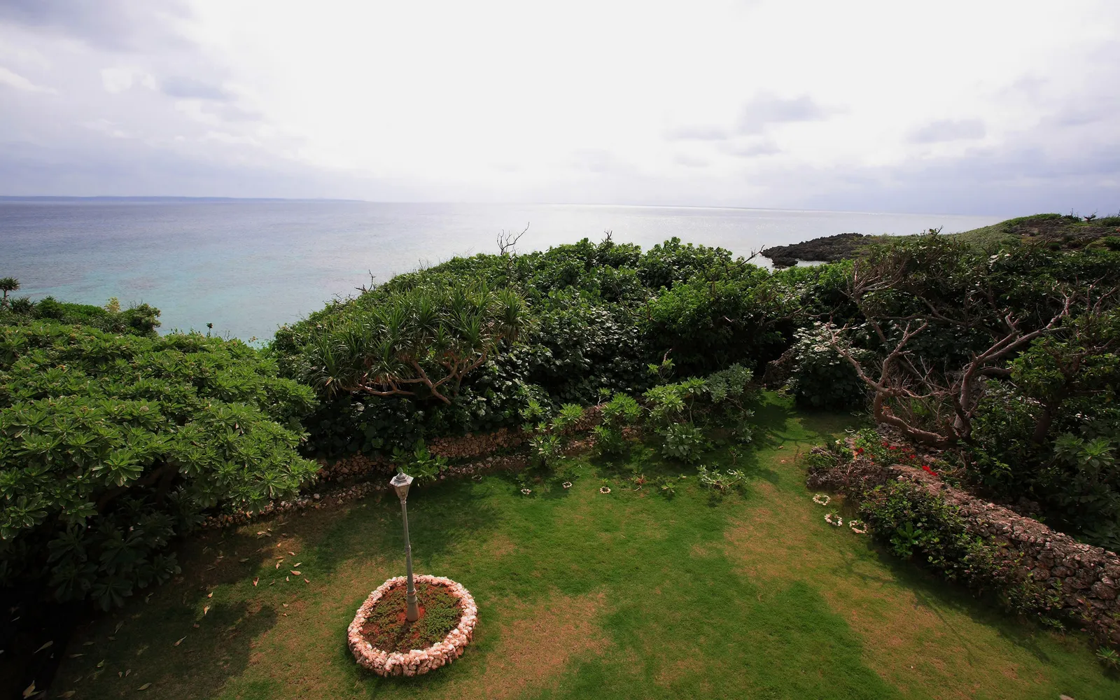 A view of a green garden with a stone wall and a lamp post, overlooking the blue ocean under a cloudy sky.