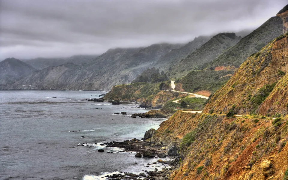A winding road hugs a steep, vegetated cliff overlooking the ocean on a cloudy day.