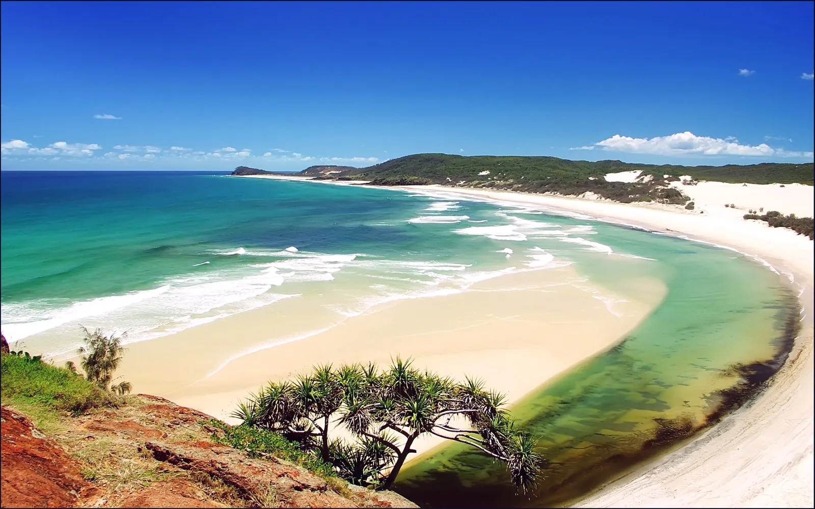 A sweeping view of a tropical coastline with turquoise ocean waves crashing on a white sand beach, backed by green hills and a clear blue sky.