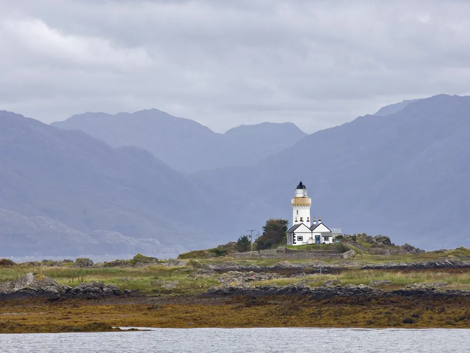 A white lighthouse with a black top stands on a rocky island with green grass and distant mountains under a cloudy sky.