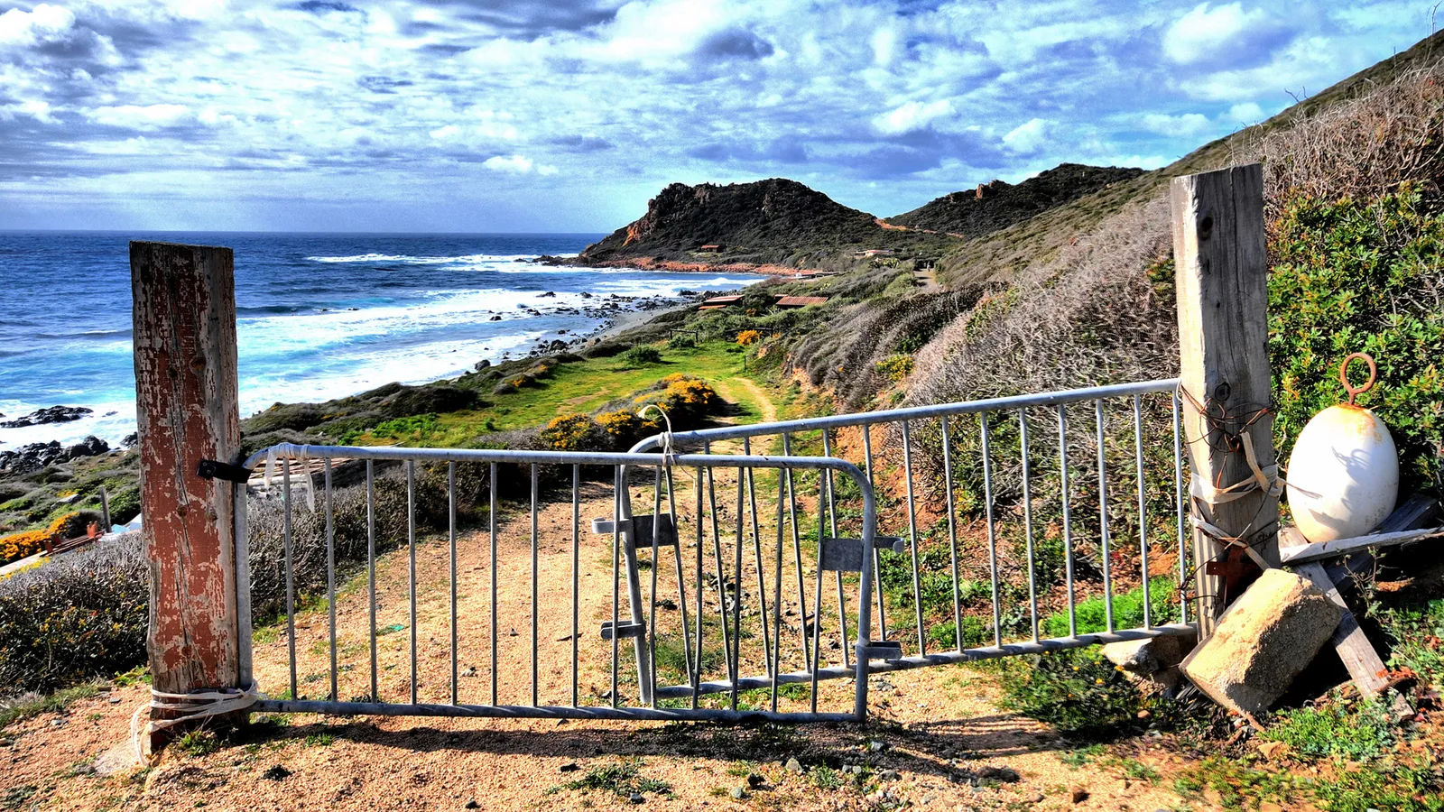 A metal gate stands open on a dirt path leading towards a rocky coastline and the blue ocean.
