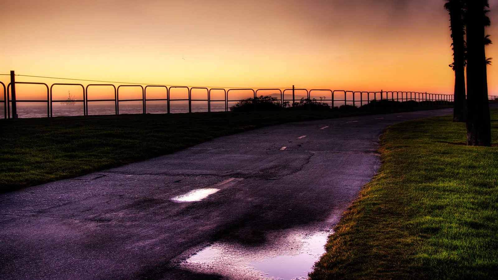 A paved path curves along a grassy coastline with a fence overlooking the ocean at sunset.