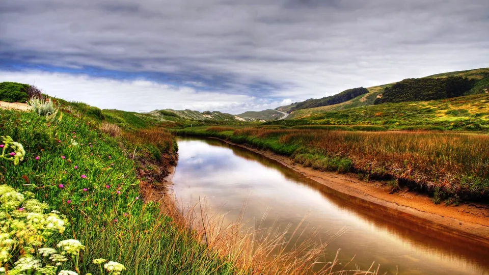 A calm river flows through a landscape of green hills and grassy banks under a cloudy sky.