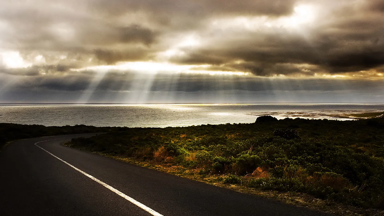 A winding asphalt road leads towards a dramatic ocean under a sky with sunbeams breaking through dark clouds.