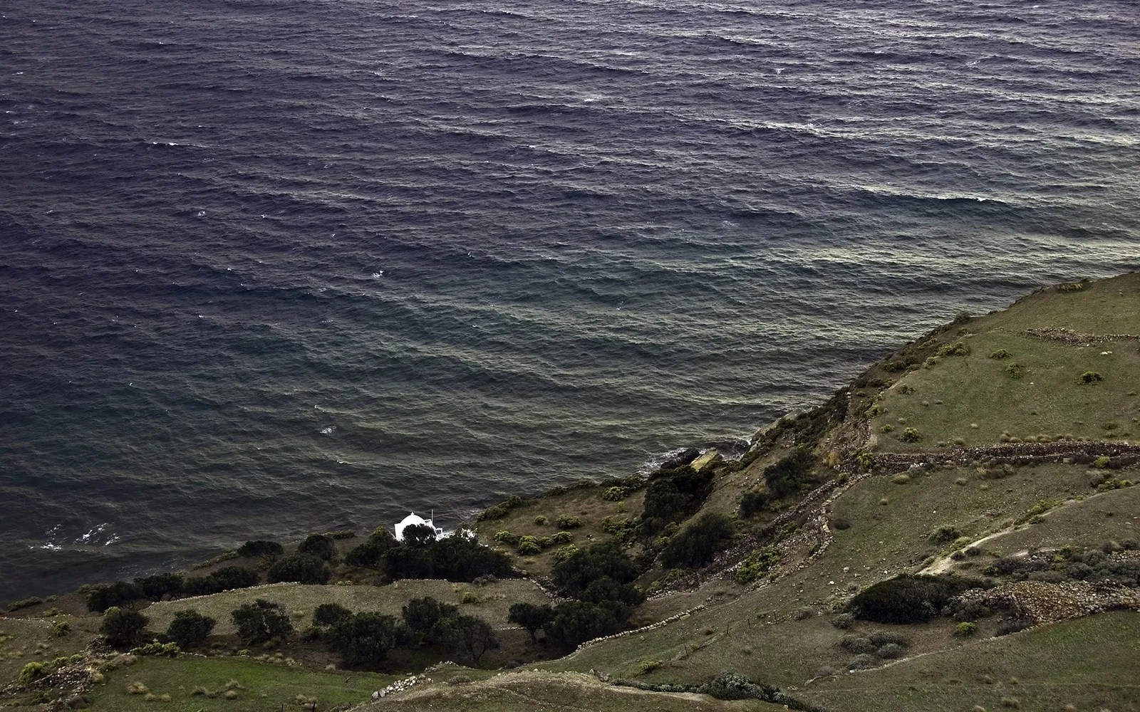 A view from a grassy cliff overlooking a dark, choppy sea with a small white building nestled on the shore.