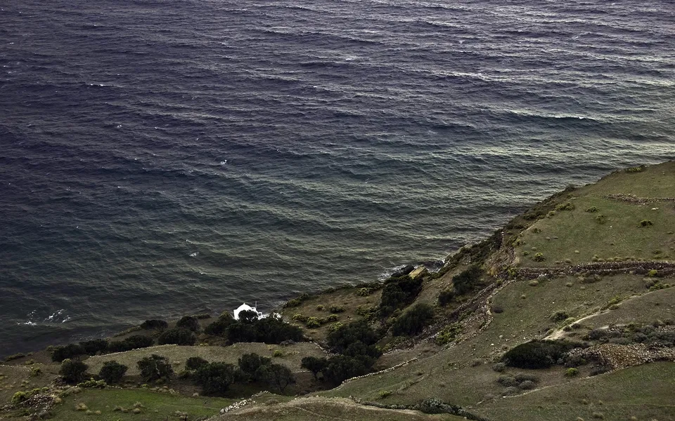 A view from a grassy cliff overlooking a dark, choppy sea with a small white building nestled on the shore.