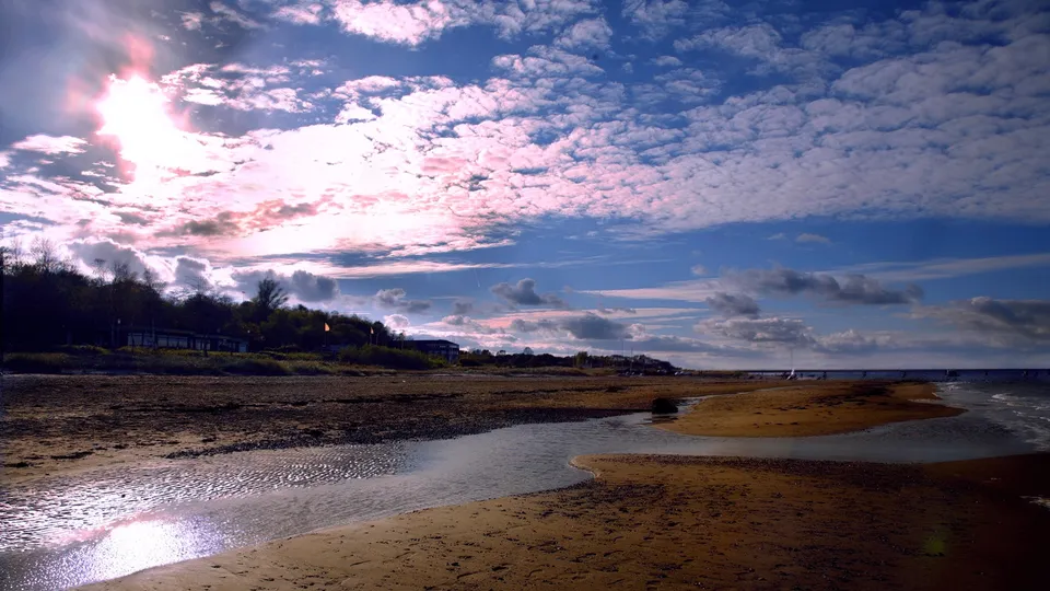 A wide shot of a sandy beach with a small stream flowing towards the ocean under a dramatic sky.