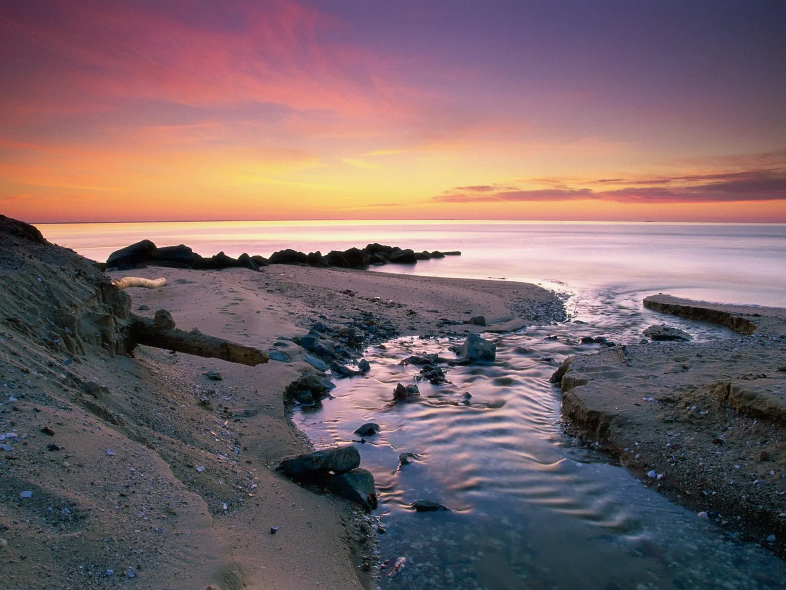 A small stream flows across a sandy beach towards the ocean during a colorful sunset.
