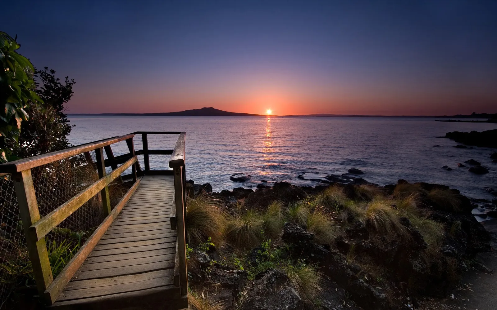 A wooden walkway leads to a viewpoint overlooking the ocean at sunrise, with a distant island on the horizon.