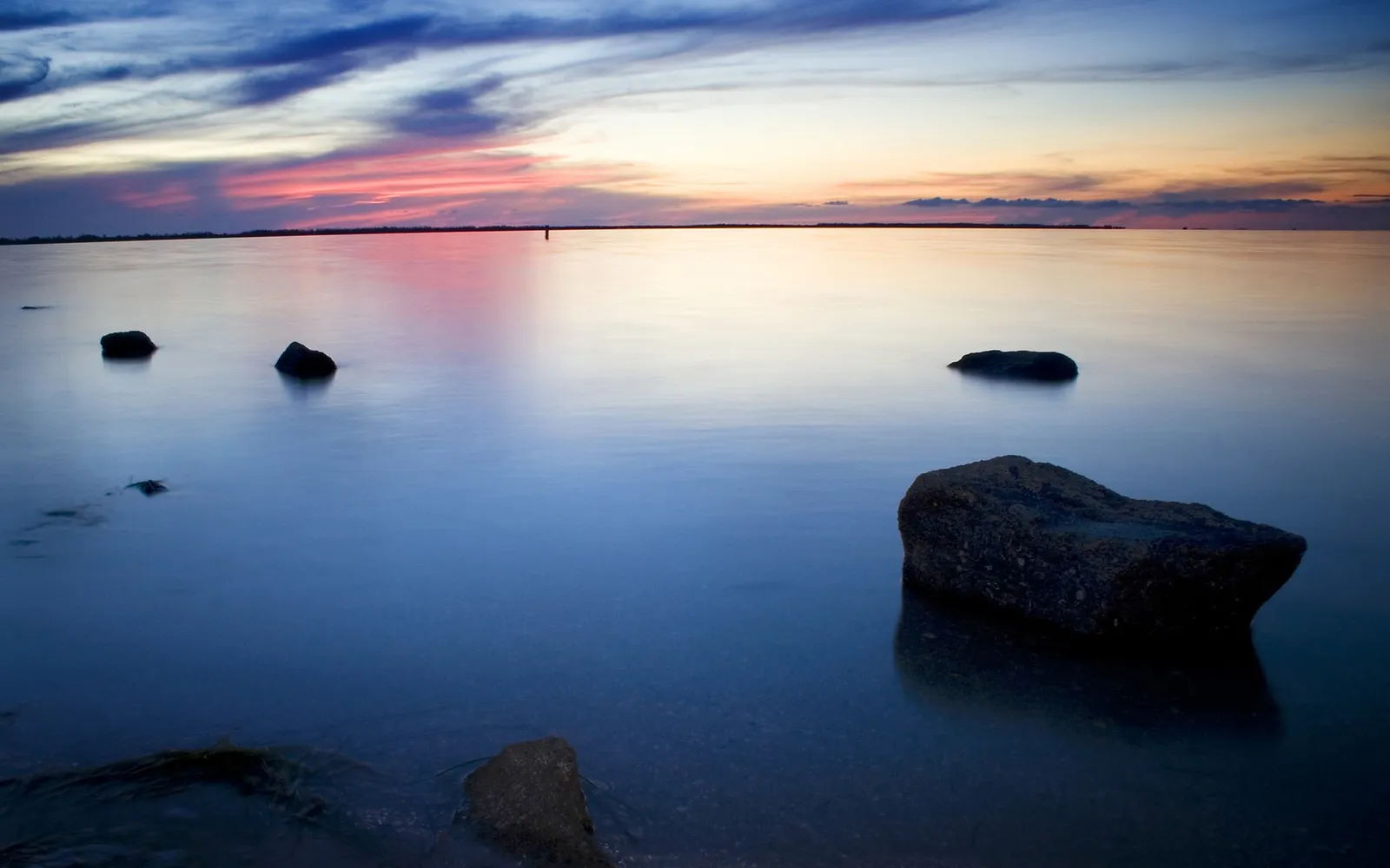A serene coastal sunset with pink and blue clouds reflected in the calm water, with several dark rocks visible.