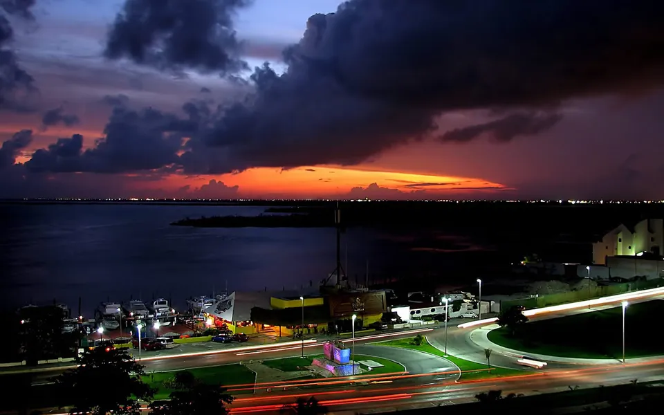 A dramatic sunset with dark clouds over a coastal city with lights and car trails.