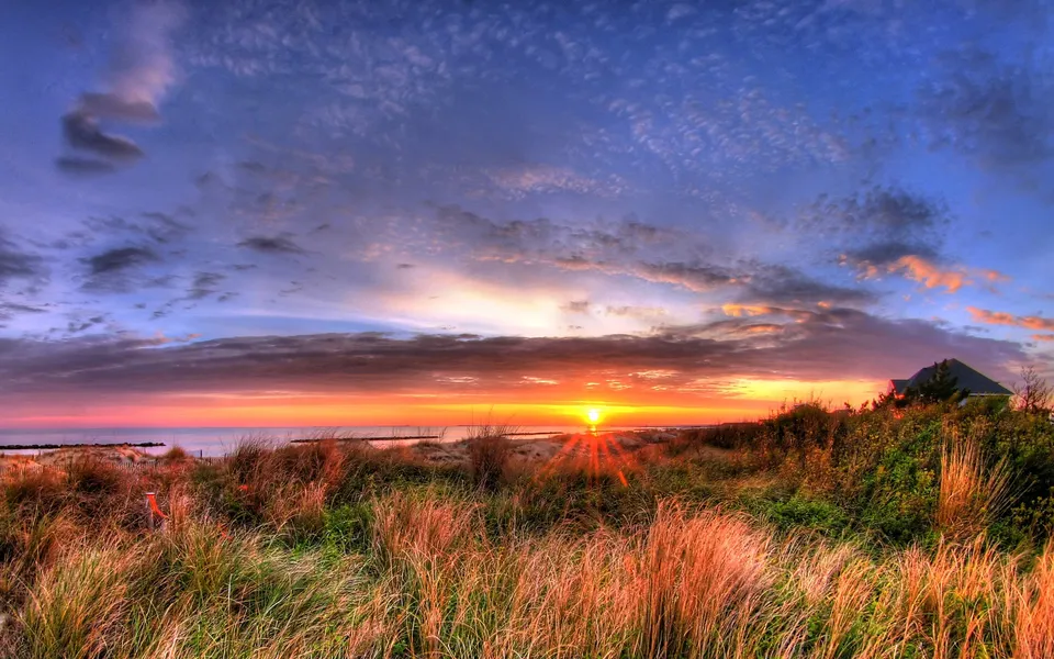 A vibrant sunset over the ocean with silhouetted dune grass in the foreground and a house on the right.