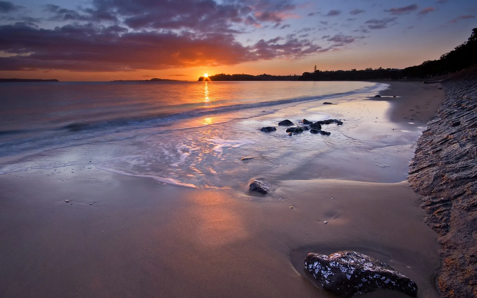 A vibrant sunset casts golden and purple hues over a calm beach with gentle waves lapping the shore.