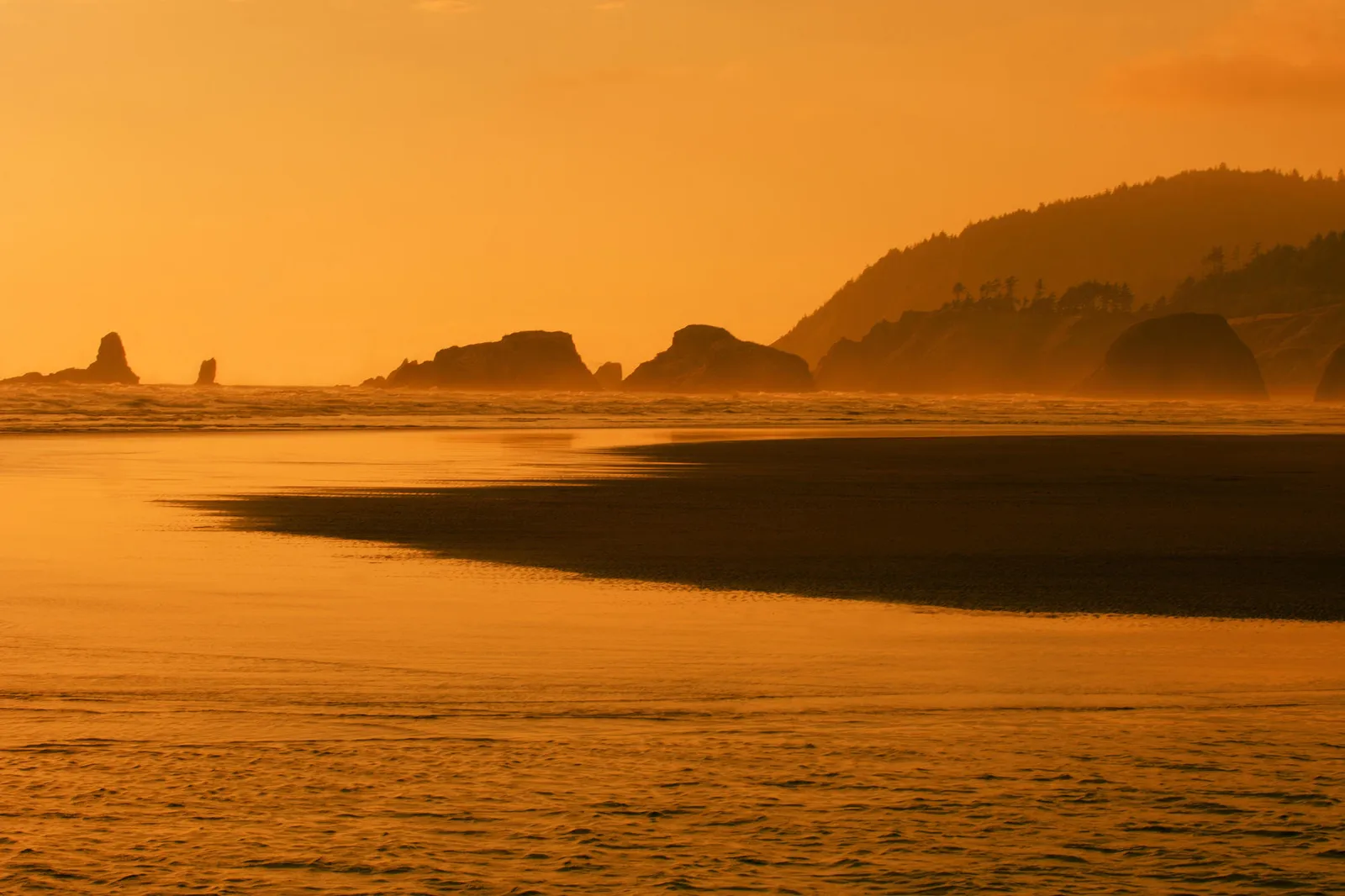 A wide shot of a beach at sunset with the sky and ocean glowing orange, silhouetting coastal rocks and hills.