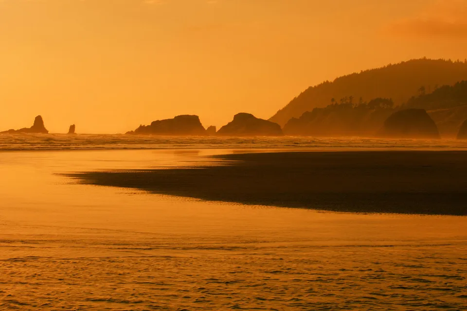 A wide shot of a beach at sunset with the sky and ocean glowing orange, silhouetting coastal rocks and hills.