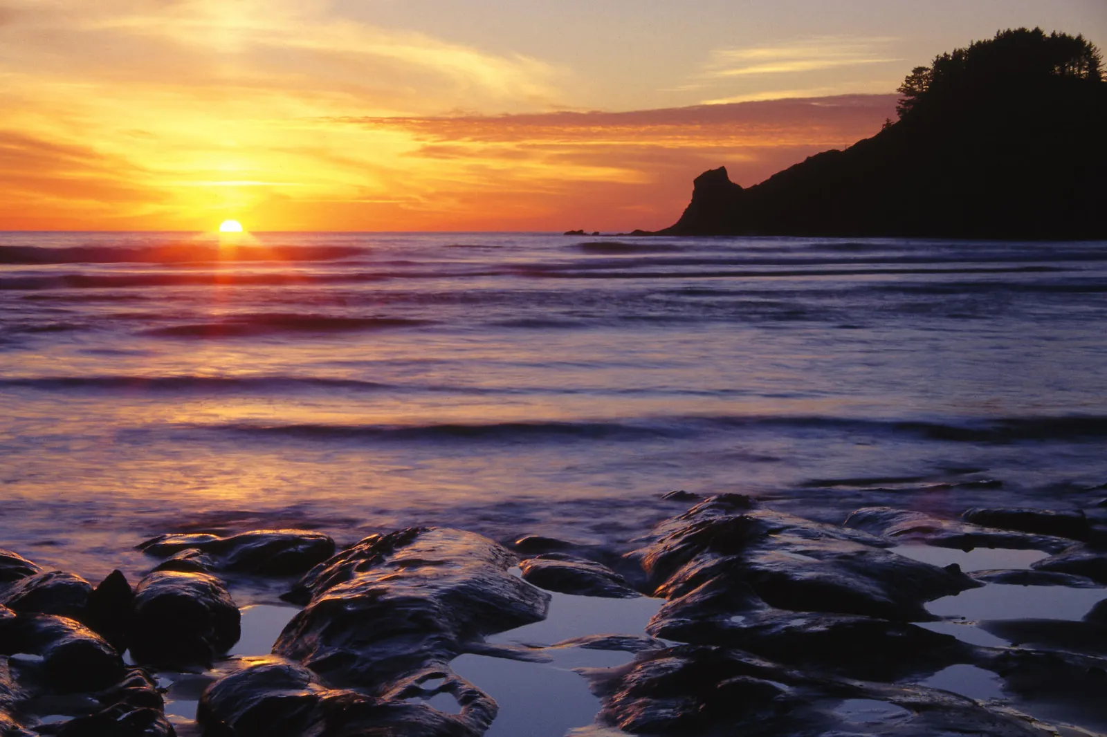 A vibrant sunset over the ocean, with silhouetted cliffs and rocks in the foreground.