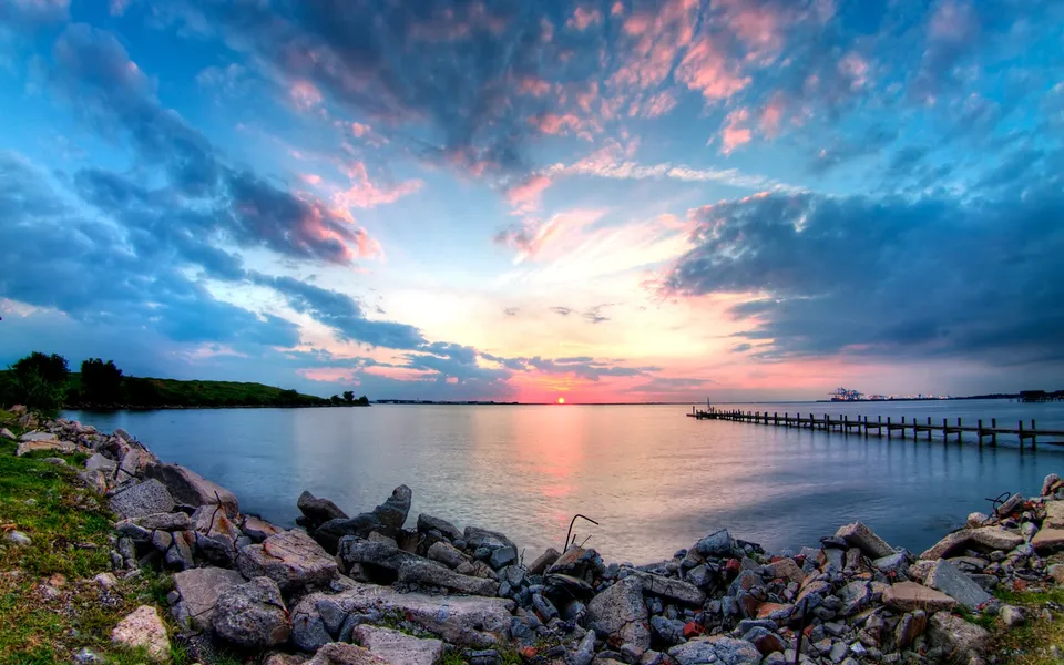 A panoramic view of a sunset over calm coastal waters, with a pier extending into the sea and a foreground of rocks.