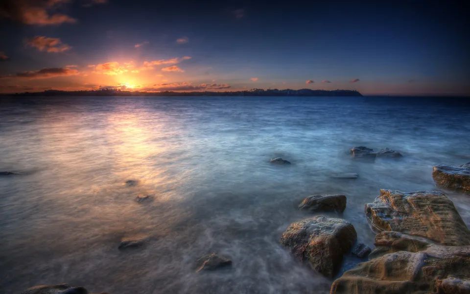 A vibrant sunset casts golden light across the ocean, with silhouetted rocks in the foreground.
