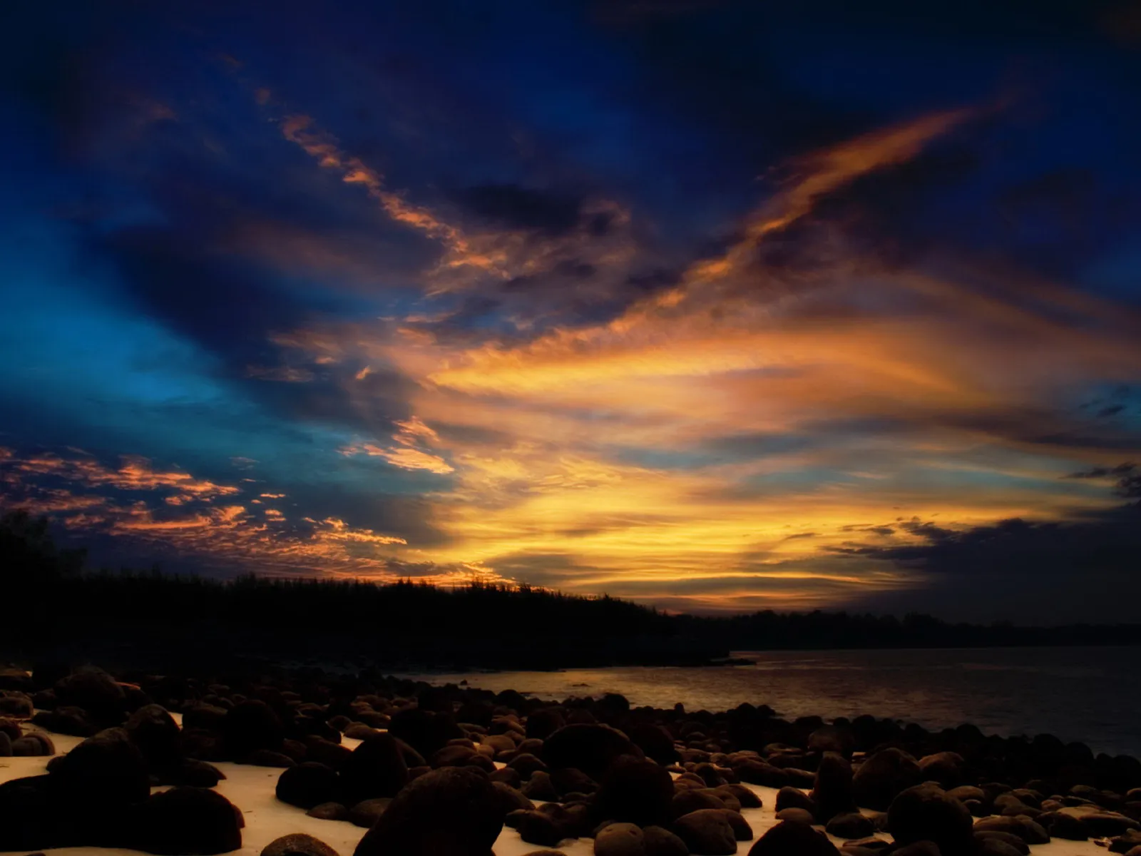 A dramatic sunset with vibrant orange and blue clouds over a rocky beach and calm water.