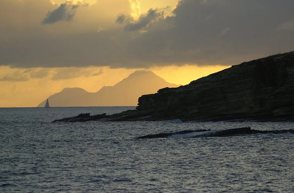 A sailboat is visible on the ocean in front of silhouetted mountains during a cloudy sunset.