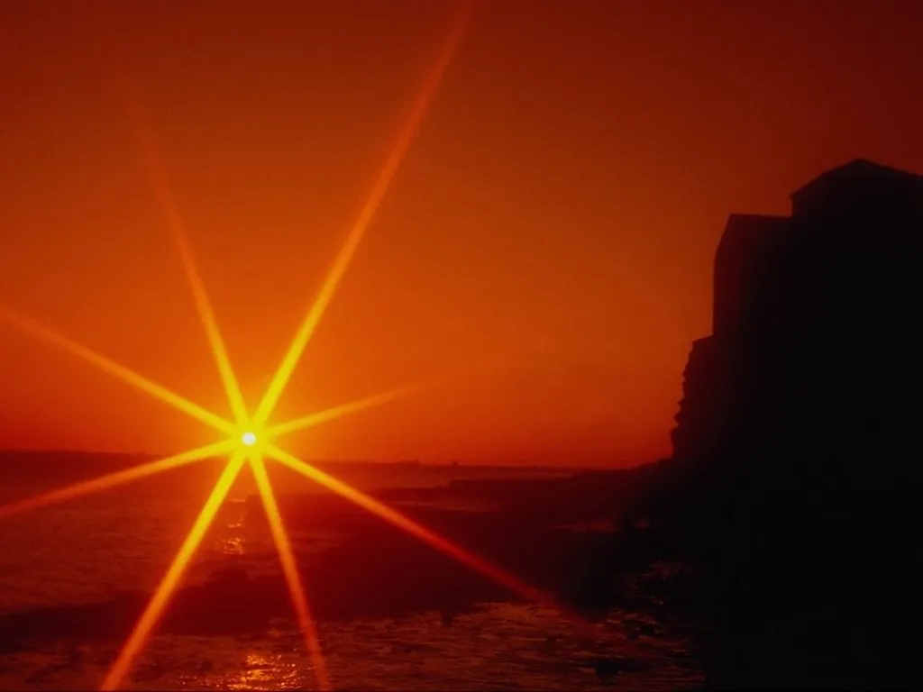 A bright sunburst illuminates the sky over the ocean, with dark silhouettes of buildings on a cliff.