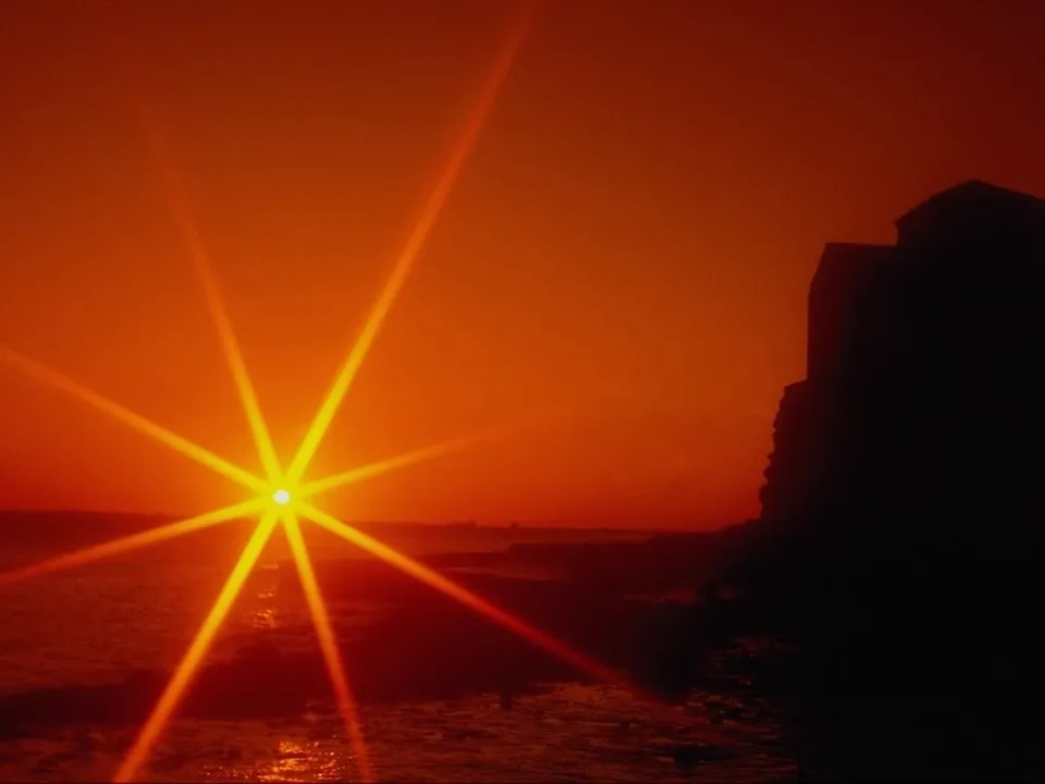 A bright sunburst illuminates the sky over the ocean, with dark silhouettes of buildings on a cliff.