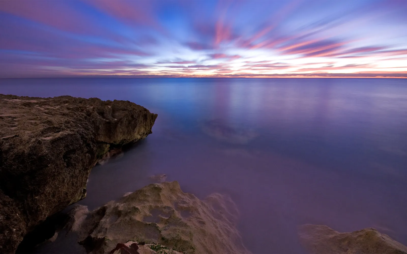 A long exposure shot of a dramatic sunset over the ocean, with streaks of pink and blue in the sky and rocky shore in the foreground.