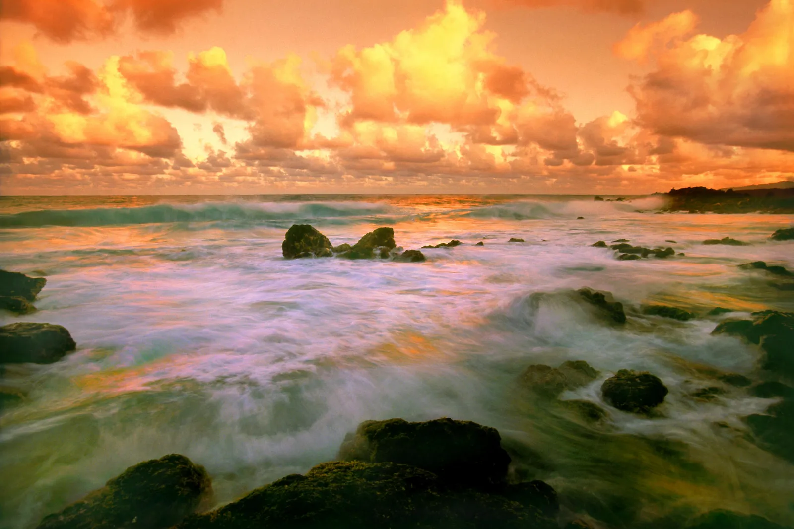 Waves crash on rocks along the coast during a vibrant sunset with orange clouds.