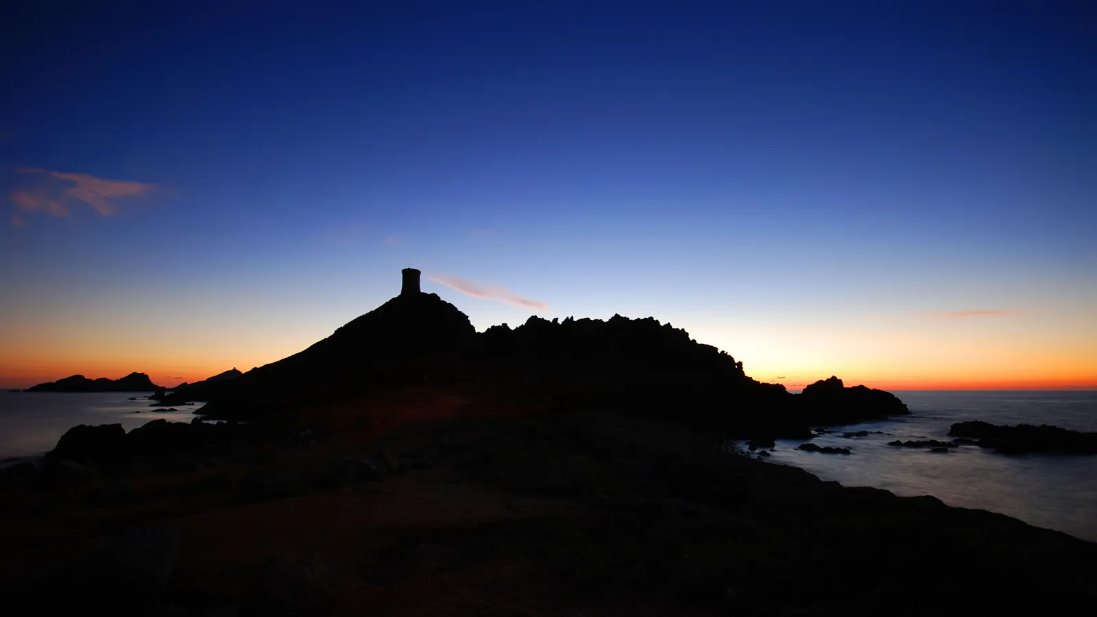 Silhouette of a tower on a rocky island against a gradient blue and orange sky at dusk.