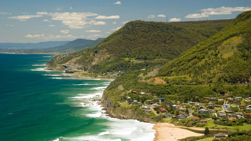 An aerial view of a coastal town with houses nestled on a green hillside overlooking the ocean and a sandy beach.