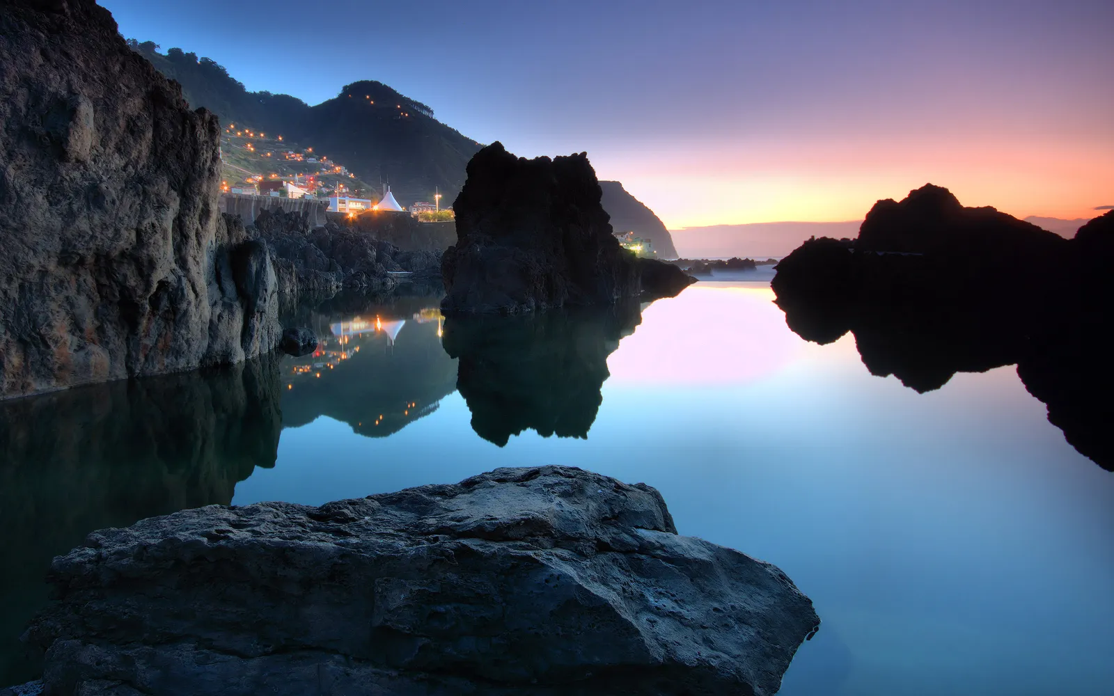 A serene coastal scene at twilight, with calm water reflecting the sky and distant village lights on a mountainside.