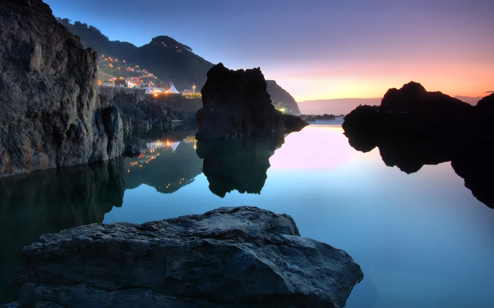 A serene coastal scene at twilight, with calm water reflecting the sky and distant village lights on a mountainside.