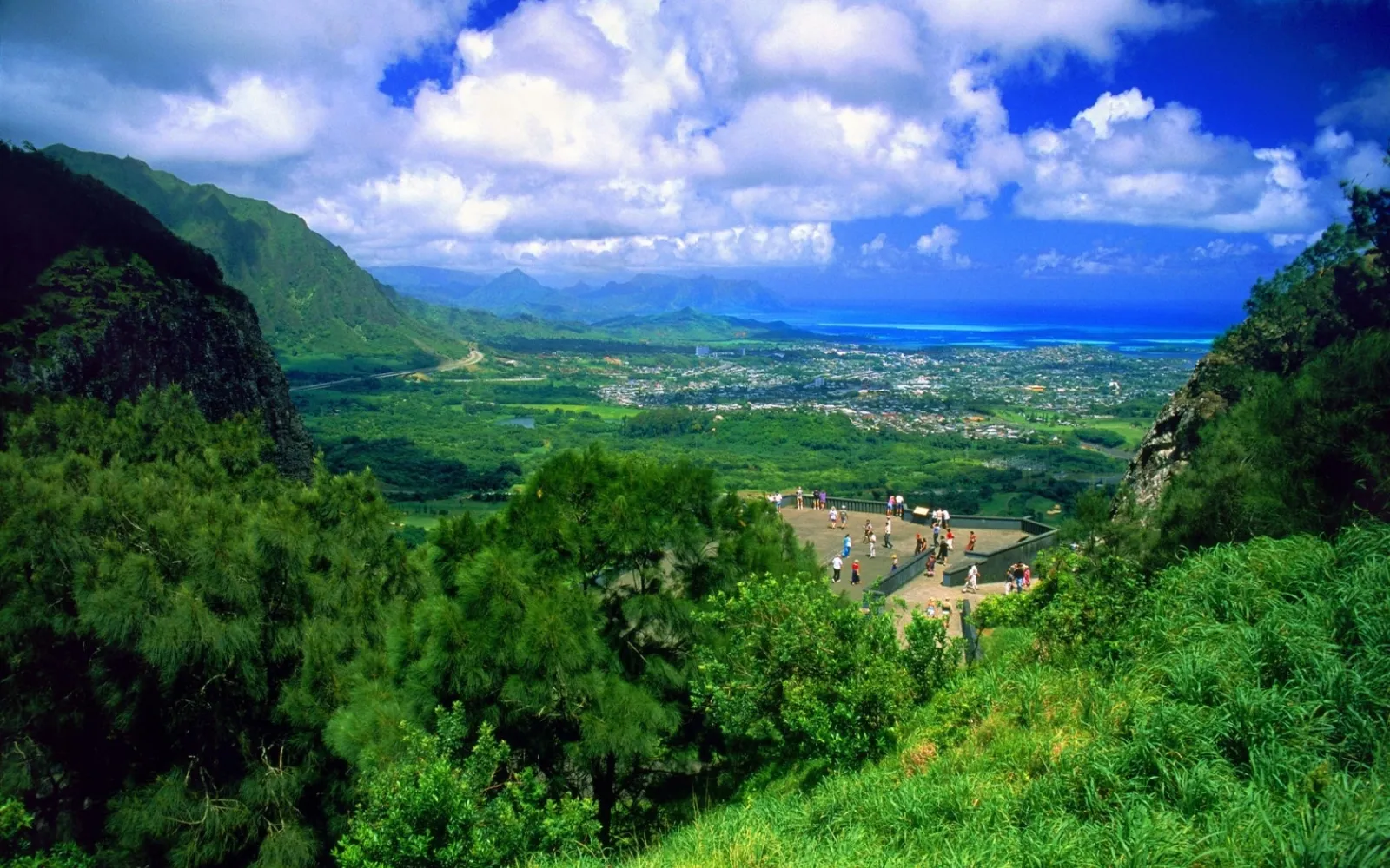 A scenic overlook offers a view of a green valley leading to a coastal city and the blue ocean under a cloudy sky.