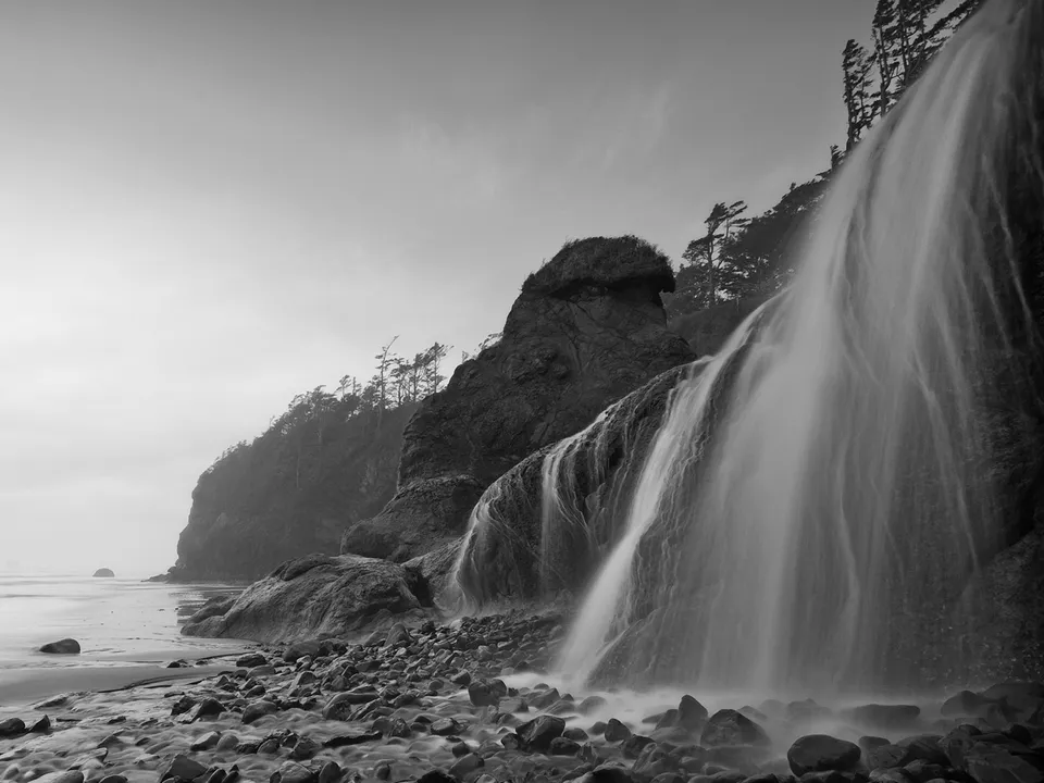 A black and white image of a waterfall flowing down a cliff onto a rocky beach with the ocean in the background.