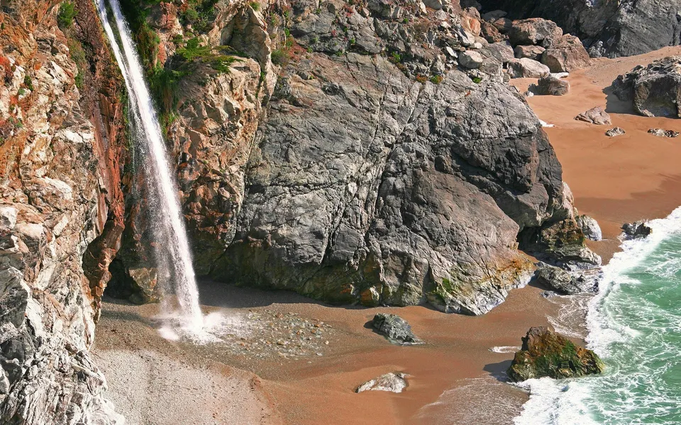 A tall waterfall plunges from a rocky cliff onto a sandy beach next to the ocean.