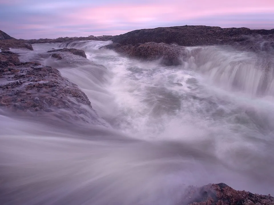 A long exposure captures the soft flow of water over dark, textured rocks under a pastel sky.