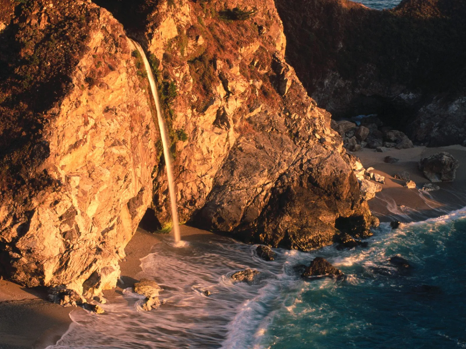 A thin waterfall cascades down a rocky cliff face onto a sandy beach, with waves crashing on the shore.