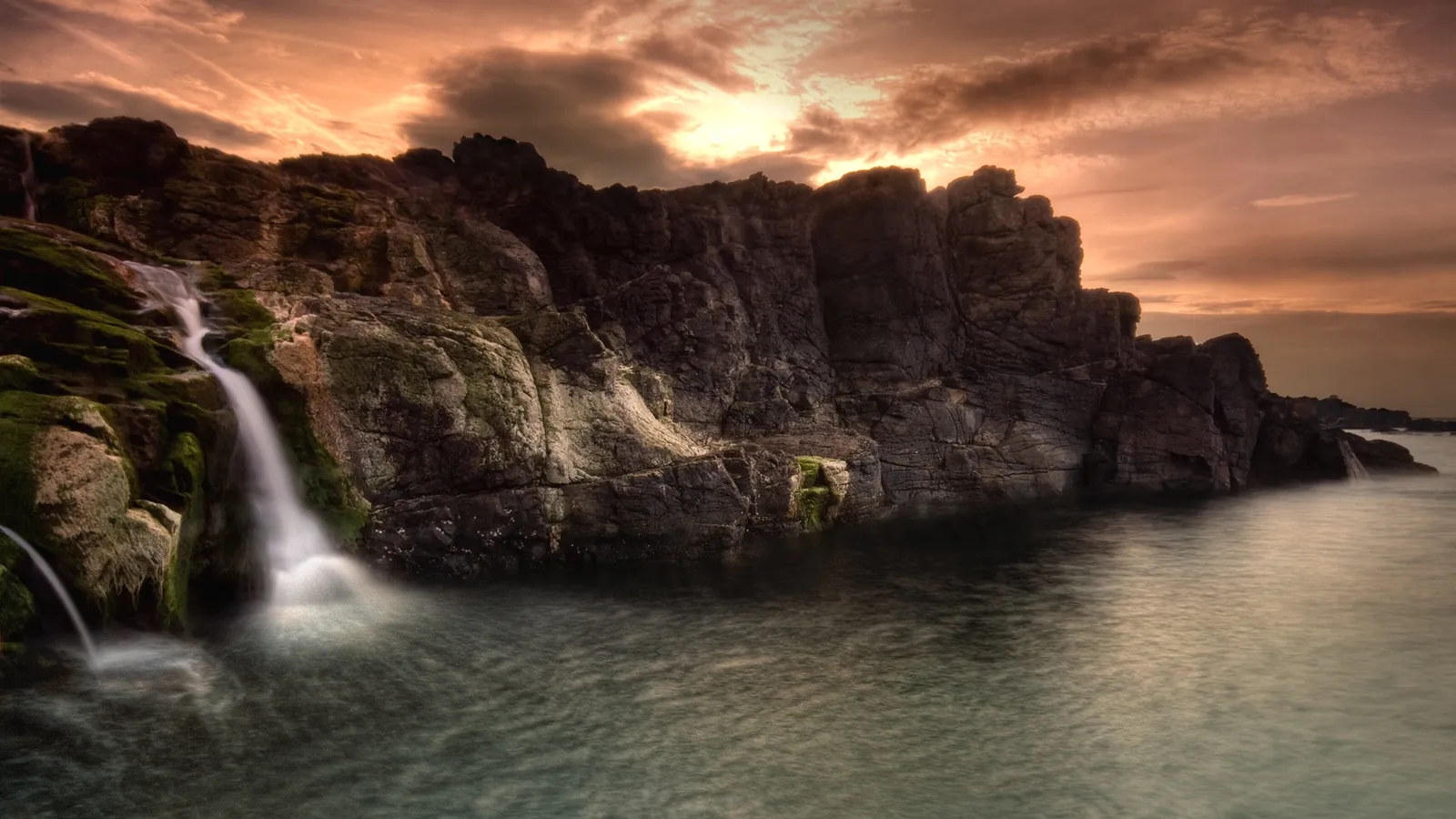 A small waterfall cascades down mossy rocks into the sea under a dramatic sunset sky.