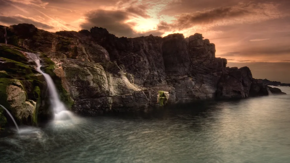 A small waterfall cascades down mossy rocks into the sea under a dramatic sunset sky.