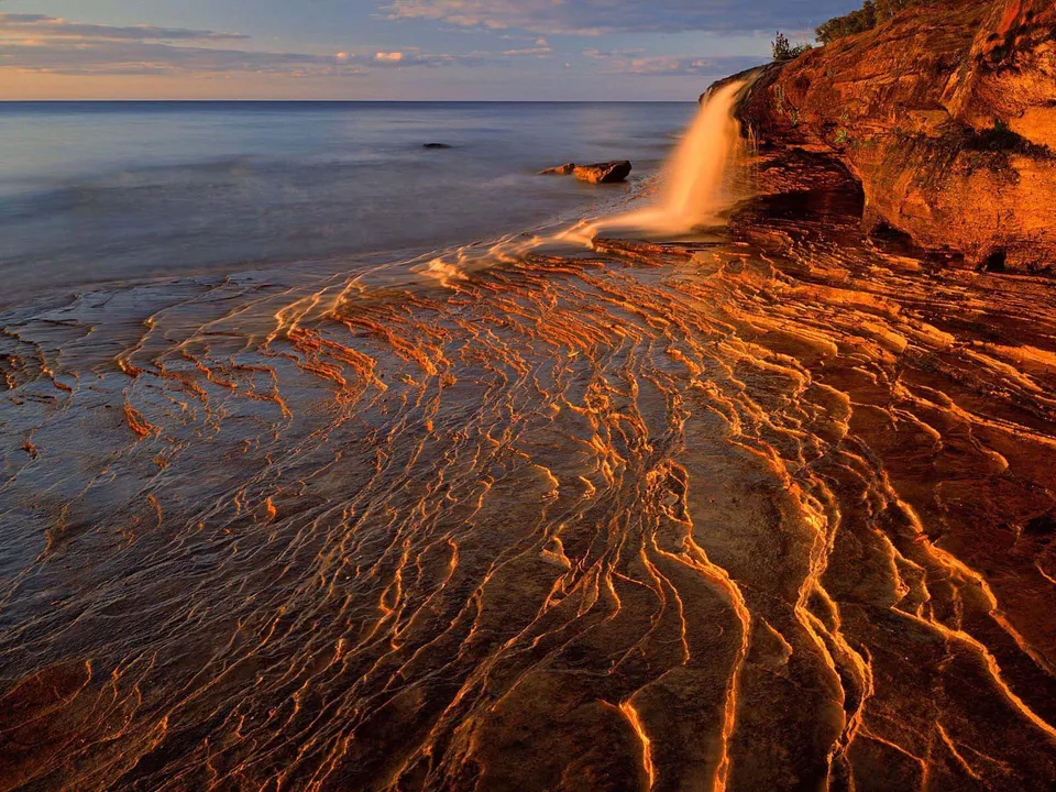 A waterfall flows over a rocky cliff onto a textured shoreline with patterns carved by water.