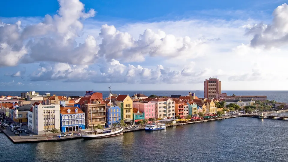 A row of colorful buildings line a waterfront with boats docked and the ocean in the background.