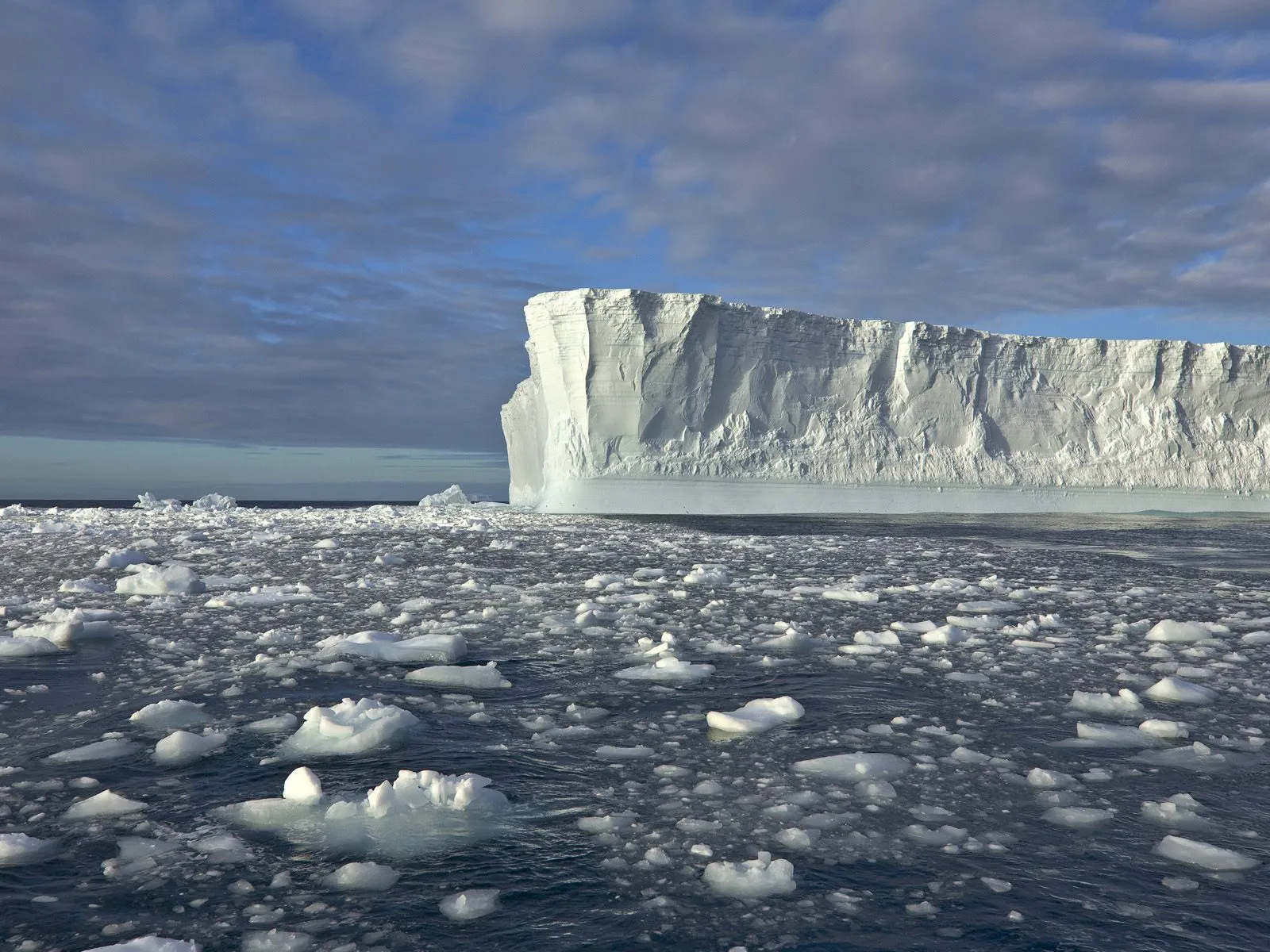A vast, sheer-faced iceberg floats in the ocean surrounded by smaller ice chunks.