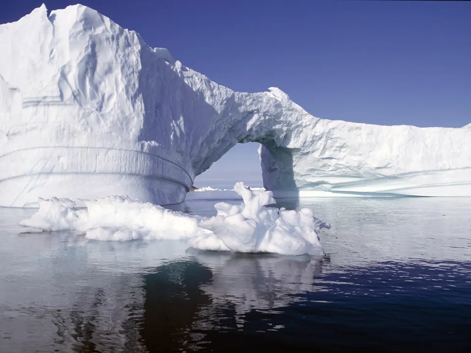 A colossal iceberg forms a natural archway, with calm water reflecting the ice and the clear blue sky.